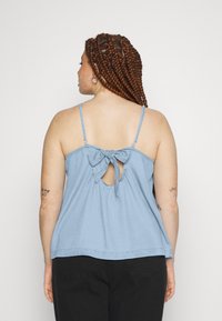Top sem mangas azul claro com costas em forma de buraco, detalhe de laço amarrado e um corte descontraído. Feito de um tecido suave com uma textura lisa.