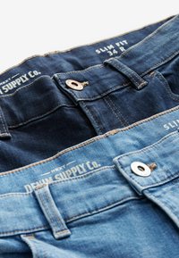 Două perechi de blugi slim fit în nuanțe de albastru închis și deschis. Caracteristicile includ accesorii din cupru, detalii cusute și etichete pe banda de talie.