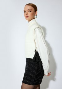 Weißer strukturierter Pullover über einer langärmeligen weißen Bluse, kombiniert mit einem schwarzen Rock mit vertikalen Streifen und einem Ripsband zum Binden in der Taille.