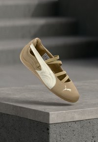 Μπεζ και λευκό μπαλέτο Puma με τρεις ελαστικές λωρίδες, αιωρούμενο πάνω από μια γκρίζα τσιμεντένια άκρη με θολά σκαλοπάτια στο φόντο.