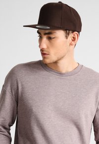 Brun, tætsiddende baseballcap med flad skygge, lavet af struktureret stof; har et cirkulært logo-patch foran og ventilationshuller.
