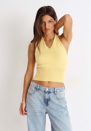 Mujer con el pelo largo y castaño lleva un top amarillo sin mangas y vaqueros azul claro, de pie con una mano detrás de la cabeza frente a un fondo liso.