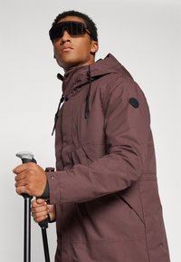 Veste isolante brune avec capuche, accents noirs et cordons de serrage. Le modèle tient des bâtons de trekking et porte des lunettes de soleil noires. Tissu texturé.