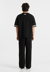 Juodas medvilninis marškinėliai su trumpomis rankovėmis, su ryškiais kontrastingais raštiniais akcentais ant rankovių, derinami su juodomis plačiomis kelnėmis ir baltais sportbačiais.