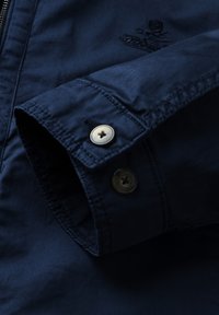 Poignet de chemise bleu marine avec deux boutons blancs, couture détaillée et logo brodé partiellement visible sur le tissu.