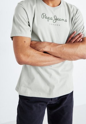 Osoba ze skrzyżowanymi ramionami, ubrana w jasnoszarą koszulkę "Pepe Jeans London" i ciemne spodnie, kadrowana od ramion do bioder.