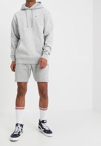 Felpa grigia con tasca frontale e stringhe regolabili, abbinata a pantaloni corti grigi. Calzini bianchi con strisce rosse e scarpe da ginnastica blu navy.