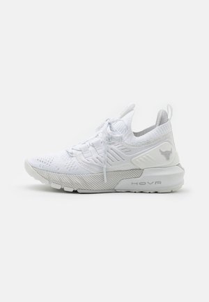 Zapatilla deportiva blanca con parte superior de malla, diseño con cordones, suela gruesa etiquetada "HOVR," y logotipo de cabeza de toro en el talón. Vista lateral.