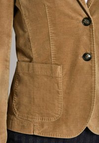 Tannfarbene Cordjacke mit zwei dunklen Knöpfen und einer großen quadratischen Fronttasche, getragen über dunklen Hosen.