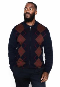 Námornícky zipsový sveter s kosoštvorcovým vzorom hnedých a červených farieb, s golierom a textúrovanou tkaninou, priliehavý na bielom pozadí.