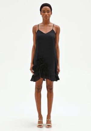 Femme portant une robe noire sans manches avec des fines bretelles et trois ornements en fleurs de tissu près de l'ourlet, assortie à des sandales dorées.