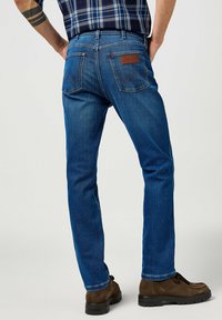 Vaqueros de denim azul con un diseño de pierna recta y parche de cuero en la marca. Cuenta con dos bolsillos traseros y sutiles detalles de costura. Zapatos marrones visibles.