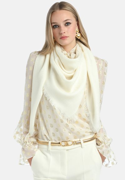 Baltas šalikas, pagamintas iš minkštos medžiagos, su pūstų kraštu. Dėvimas ant raštuoto bluzono su šviesiais aukso akcentais.