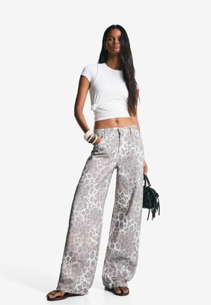 Leopardprint wijde broek van zacht materiaal met lichtgrijze en donkere vlekken. Gepaarde met een witte nauwsluitende top en een zwarte handtas.