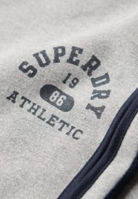 Szary materiał posiada logo "SUPERDRY", a także napisy "19", "86" i "ATHLETIC", podkreślone ciemnoniebieskim wykończeniem na brzegach.