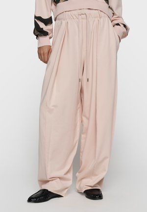 Mujer que lleva pantalones anchos sueltos de color rosa pálido con cintura de cordón y zapatos negros sin cordones, manos en los bolsillos.