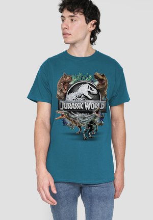 Junger Mann trägt ein blaues T-Shirt mit Jurassic World-Logo und Bildern von brüllenden Dinosauriern vorne und schaut zur Seite.