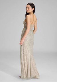 Długa, dopasowana sukienka wieczorowa z głębokim dekoltem na plecach, wykonana z lśniącego beżowego materiału ozdobionego srebrnymi cekinami w zakrzywionym wzorze.