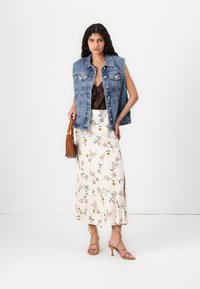 Denim ärmlös jacka över en brun topp, i kombination med en blommönstrad midiklänning och beiga klackar med remmar. Brun handväska som accent.