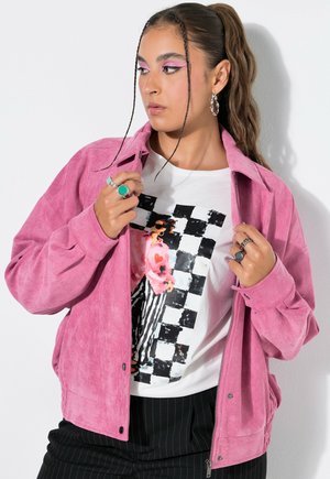 Kvinde med flettet hår bærer pink ruskindjakke over en grafisk hvid t-shirt med sort og pink ternet design, sorte snorede bukser og sølvringe.