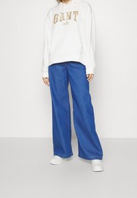 Sudadera blanca con capucha y el logo beige "GANT 1949", combinada con pantalones de mezclilla azul de pierna ancha y zapatillas blancas.
