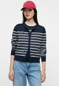 Σκούρο μπλε ριγέ κάρντιγκαν με λευκές οριζόντιες ρίγες, στρογγυλή λαιμόκοψη, κουμπί μπροστά και χαλαρή εφαρμογή, συνδυασμένο με ροζ καπέλο μπέιζμπολ.