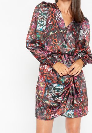 Femme portant une robe portefeuille à motifs multicolores avec des manches longues bouffantes et des poignets froncés, debout devant un fond blanc.