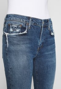 Vaqueros de mezclilla azul oscuro con parches desgastados, bordes deshilachados, diseño de cinco bolsillos y corte clásico de pierna recta.