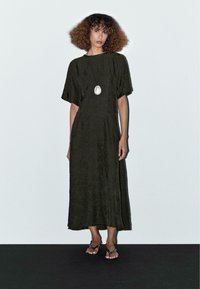 Dunkelgrünes, knöchellanges Kleid mit kurzen Ärmeln und einem strukturierten Stoff. Tragen mit einer silbernen Anhänger-Halskette und schwarzen Sandalen.