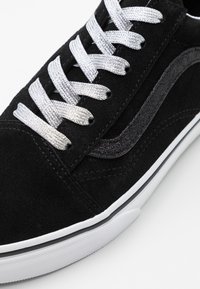 Sneaker in suede nera con suola in gomma bianca, lacci argentati e striscia laterale grigio scuro. Superficie texturizzata con dettagli di cucitura.