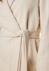 Cappotto avvolgente beige realizzato in tessuto testurizzato, con cintura da annodare e maniche larghe. Il design è semplice e classico con una finitura morbida.