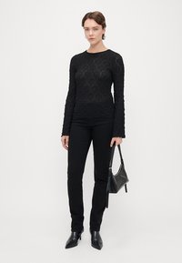 Czarny, teksturowany, długi top w połączeniu z dopasowanymi czarnymi spodniami i czarnymi botkami na obcasie, trzymający małą czarną torebkę.