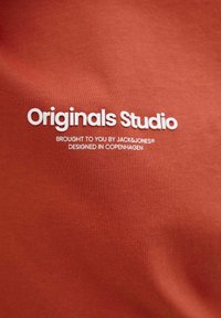 Rudos spalvos audinys su lygiu paviršiumi, su balta tekstu: "Originals Studio Brought to you by JACK&JONES Sukurta Kopenhagoje."