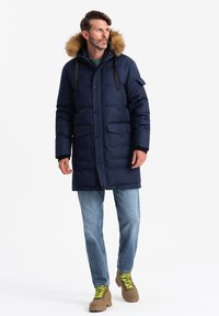 Parka blu navy con cappuccio foderato in pelliccia, tasche frontali e chiusura con zip. Indossato con jeans blu chiaro e stivali marroni con lacci verdi.