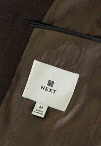 Brauner Stoff mit einem dezenten Muster, versehen mit einem weißen Etikett "NEXT", Größe "M" und der Aufschrift "MADE IN CAMBODIA". Texturiertes Innenfutter.