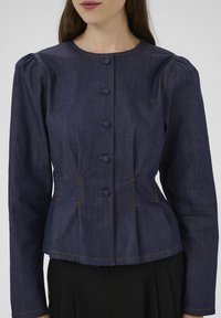 Giacca in denim scuro con maniche lunghe, scollo rotondo, chiusura con sei bottoni, spalle arricciate e dettaglio di cuciture a contrasto sull'orlo.