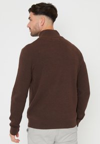 Brauner Strickpullover mit hohem Kragen, gerippten Bündchen und glatter Textur. Das Design zeichnet sich durch eine taillierte Form und minimale Details aus.