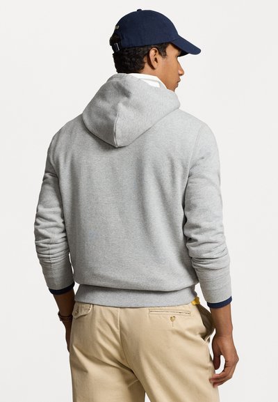 Grå bomulds-hættetrøje med hætte og ribbet manchetter, båret over beige bukser med detalje på baglommen. Kappen er navy med justerbar rem.