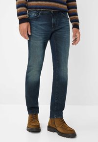 Uomo che indossa jeans blu sbiaditi, stivali in suede marrone con lacci e un maglione a righe in maglia, in piedi su uno sfondo bianco.