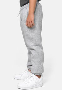Niño vestido con pantalones de chándal gris claro con puños elásticos y zapatillas blancas, de pie de lado contra un fondo liso.