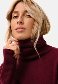 Maglione a collo alto borgogna con texture a coste e collo alto. La mano del modello è appoggiata vicino al mento, mettendo in evidenza il dettaglio del tessuto morbido e il taglio aderente.