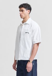 Homme portant une chemise blanche à manches courtes avec « Paternité » brodé sur la poche, jean foncé et une montre noire au poignet, regardant vers la gauche.