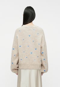 Beige knitted sweater featuring light blue heart patterns throughout. Oversized design with long sleeves, ribbed neck, and hem.
