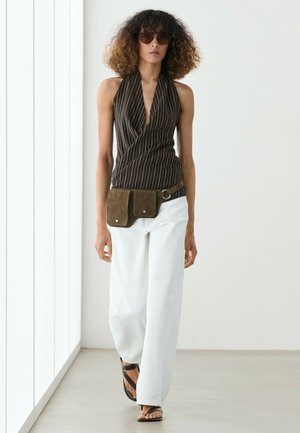 Frau mit lockigem Haar, die ein braunes gestreiftes Neckholder-Top, weiße Hose, braune Gürteltasche, schwarze Sandalen und Sonnenbrille trägt und drinnen geht.