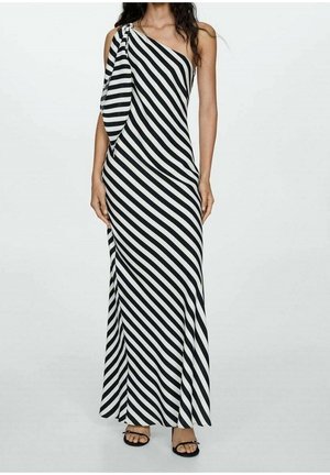 Femme portant une robe longue asymétrique rayée diagonale noire et blanche avec une manche drapée et des sandales noires ouvertes.