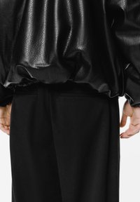 Persoon in een gestructureerde zwarte nep leren jas en zwarte broek staat met de handen relaxed aan de zijkanten tegen een eenvoudige achtergrond.