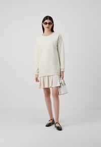 Felpa chiara e vestito crema a strati, abbinati a scarpe nere e una piccola borsa bianca. Texture morbide e forme semplici.