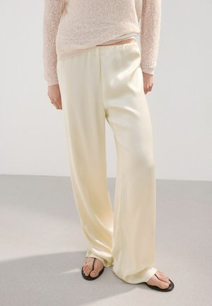 Persona vistiendo pantalones anchos de satén color crema, suéter de punto beige y sandalias planas negras, de pie sobre un suelo claro contra una pared blanca.