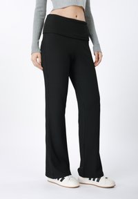 Pantalon large noir côtelé à taille haute, avec une texture douce et des coutures latérales discrètes, associé à des chaussures blanches.