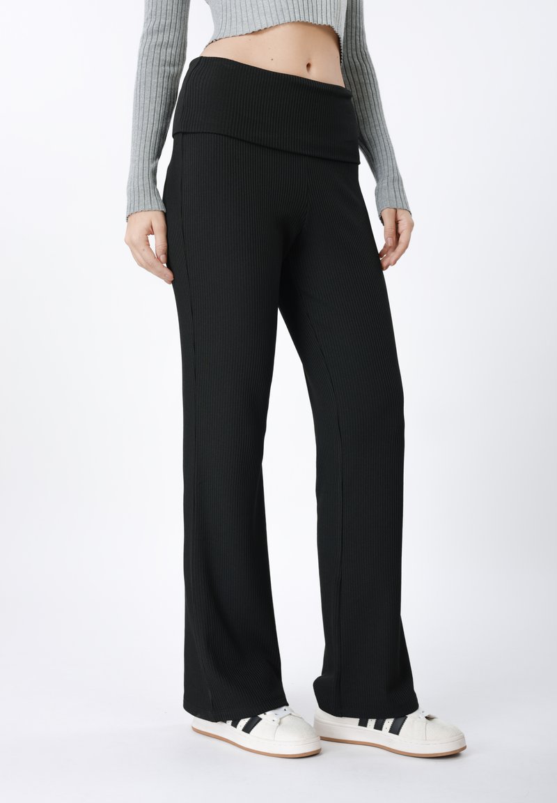 Pantalon large noir côtelé à taille haute, avec une texture douce et des coutures latérales discrètes, associé à des chaussures blanches.